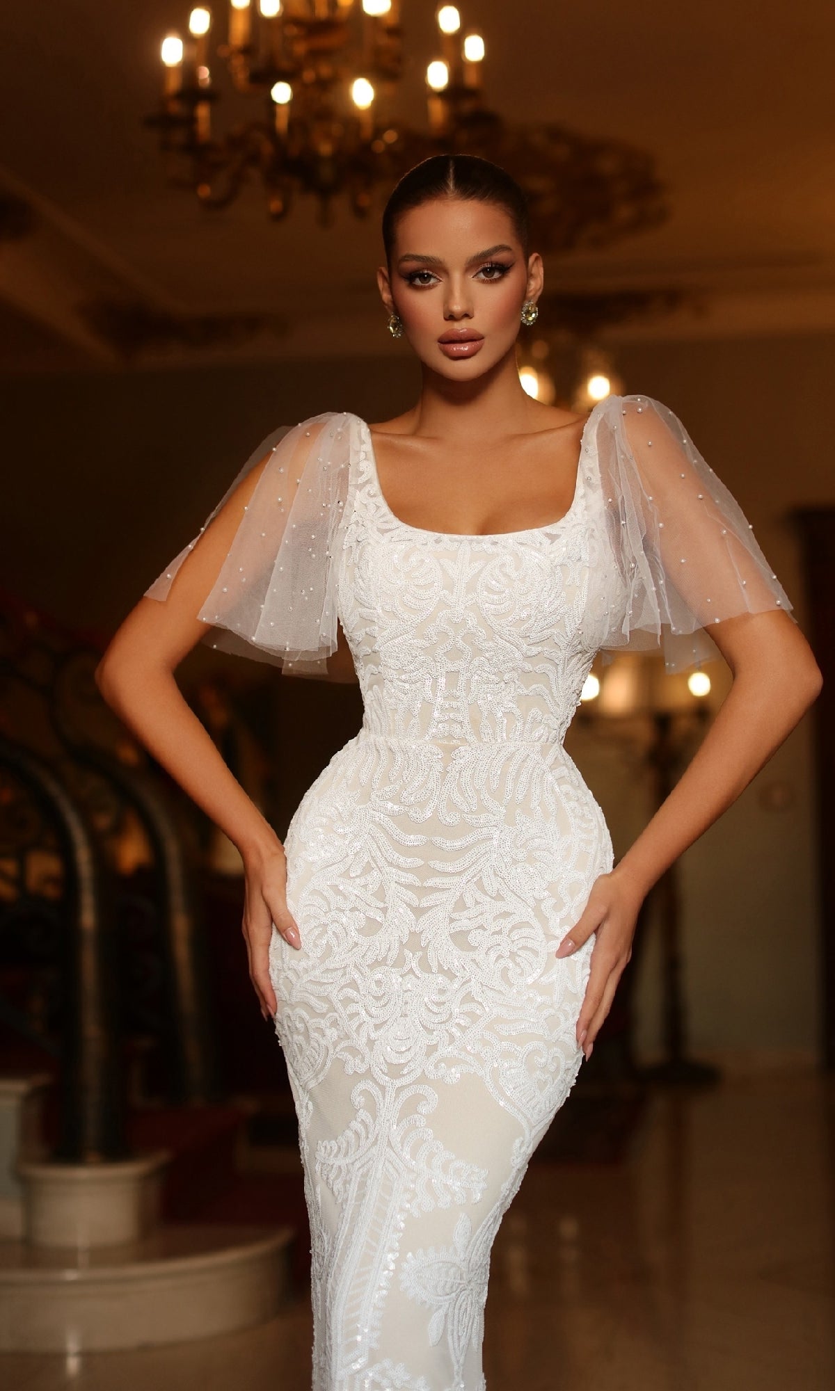 A woman poses indoors under a chandelier wearing the Tina Holly TW111, a white formal gown with short sheer puff sleeves, her hands on her hips.